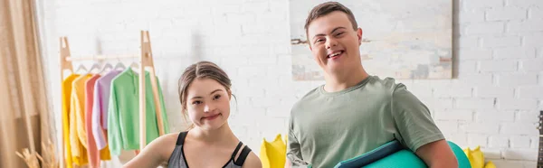 Adolescentes positivos con síndrome de Down sosteniendo alfombra de fitness en casa, pancarta - foto de stock