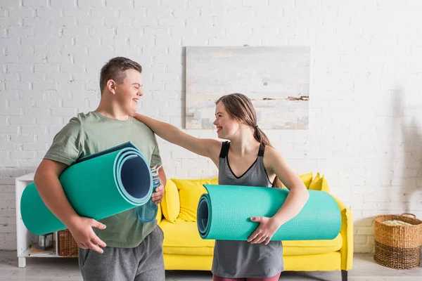 Adolescente positiva con síndrome de Down sosteniendo alfombra de fitness cerca de novio con botella de deporte en casa - foto de stock