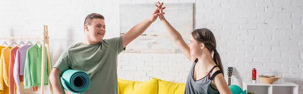 Adolescentes positivos con síndrome de Down sosteniendo alfombras de fitness y dando cinco altos en casa, pancarta - foto de stock