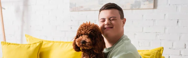 Rapaz positivo com síndrome de down fechando os olhos perto de poodle em casa, banner — Fotografia de Stock