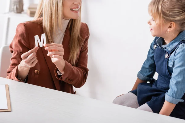 Smiling speech therapist holding letter near girl in consulting room — стоковое фото
