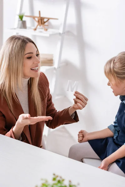 Sonriente terapeuta del habla apuntando a la carta cerca del niño en la sala de consulta — Stock Photo