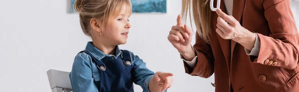 Smiling girl pointing with finger near speech therapist with letter in consulting room, banner — Stock Photo