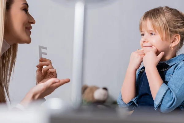 Bambino sorridente che tocca bocca vicino logopedista con lettera in sala di consultazione — Foto stock