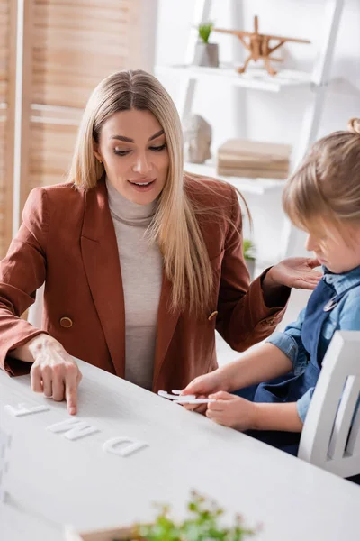 Speech therapist pointing at letter near blurred girl in classroom — Stock Photo