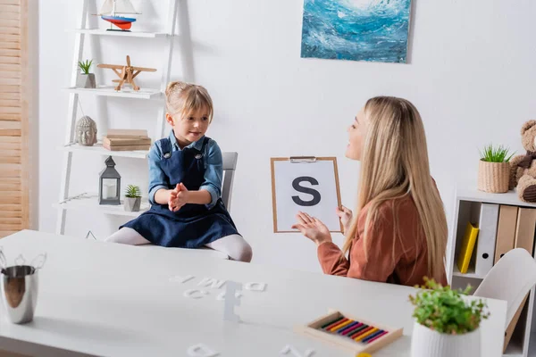 Terapeuta del habla señalando la carta en el portapapeles cerca del niño en la sala de consulta - foto de stock