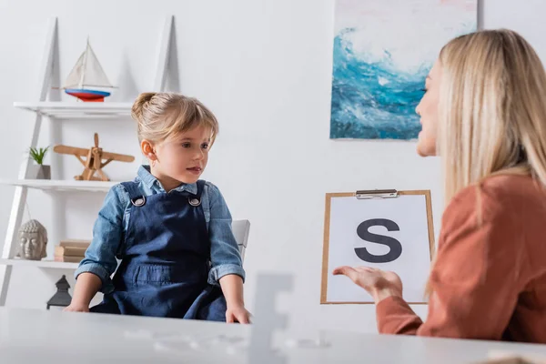 Girl sitting near blurred speech therapist with letter on clipboard in consulting room — стоковое фото