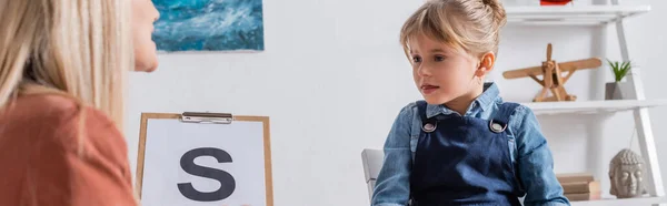 Enfant regardant un orthophoniste flou avec lettre sur presse-papiers dans la salle de consultation, bannière — Photo de stock