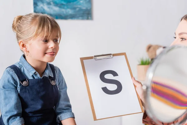 Niño sobresaliendo lengua cerca del terapeuta del habla con carta en el portapapeles en la sala de consulta - foto de stock