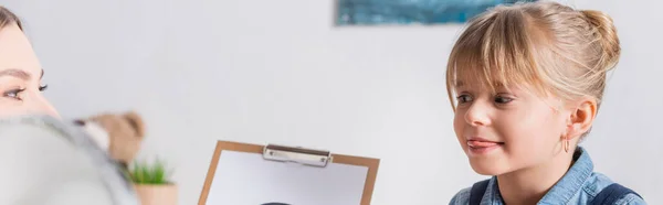 Child sticking out tongue near speech therapist with clipboard in consulting room, banner — Stock Photo