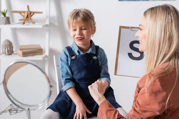 Muchacha de muecas cerca borrosa espejo y terapeuta del habla con portapapeles en la sala de consulta — Stock Photo