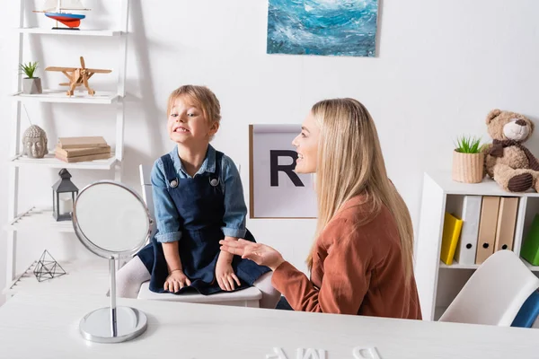 Terapeuta del habla positiva hablando y sujetando el portapapeles cerca del niño y el espejo en la sala de consulta - foto de stock