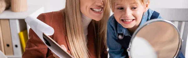 Логопед держит планшет рядом с улыбающимся ребенком и зеркало в классе, баннер — стоковое фото