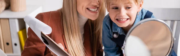 Speech therapist che tiene appunti vicino al bambino sorridente e specchio in classe, banner — Foto stock