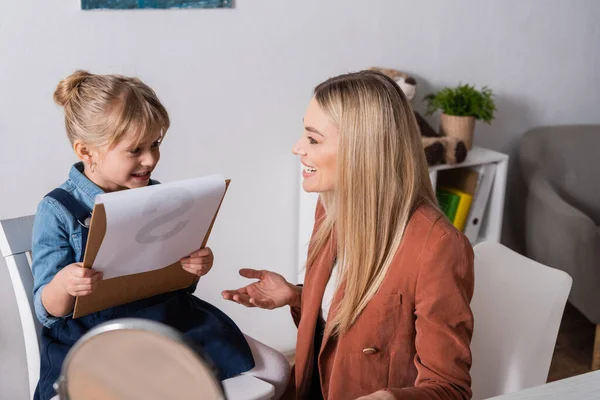 Positive speech therapist pointing with hand near girl with clipboard in classroom — стоковое фото