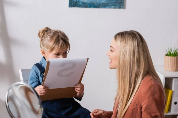 Terapeuta del habla hablando cerca de un niño sujetando portapapeles en el aula - foto de stock