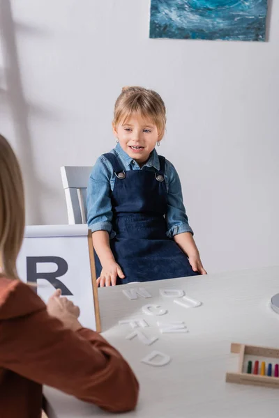 Un enfant souriant parle près d'un orthophoniste flou pointant vers une lettre sur un presse-papiers en classe — Photo de stock