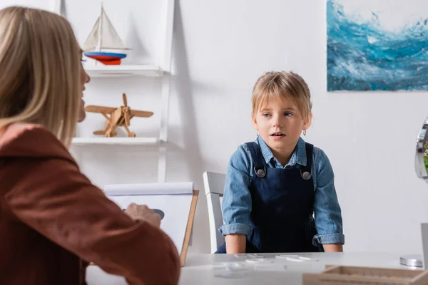 Girl talking near blurred logopedist with clipboard in consulting room — Stock Photo