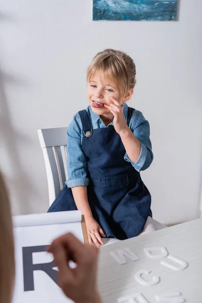 Child sticking out tongue near letters and speech therapist in classroom — Stock Photo