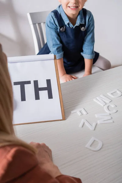 Vue recadrée d'un enfant souriant près d'un orthophoniste flou avec des lettres sur le presse-papiers en classe — Photo de stock