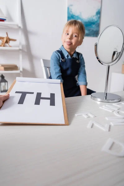 Terapeuta del habla sujetando portapapeles con letras cerca de pupilo borroso y espejo en la sala de consulta — Stock Photo