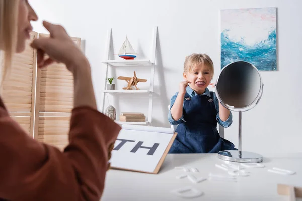 Pupil sticking out tongue and pointing with fingers while talking near mirror and blurred speech therapist in classroom — Stock Photo