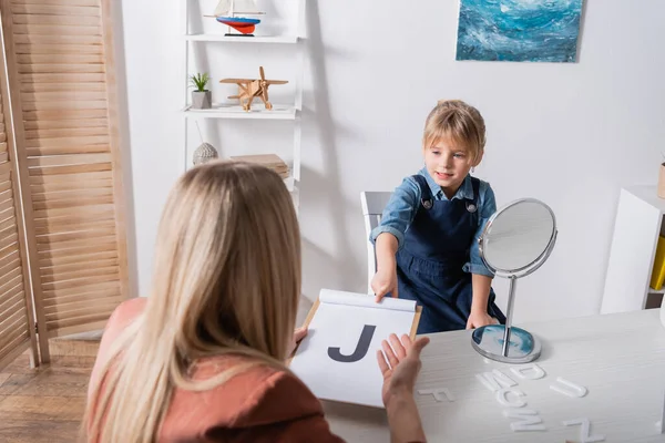 Pupil pointing at letter on clipboard near speech therapist in classroom — стоковое фото