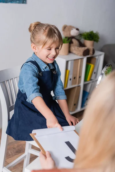 Lächelndes Kind zeigt auf Klemmbrett neben Spiegel und verschwommene Logopädin im Sprechzimmer — Stockfoto