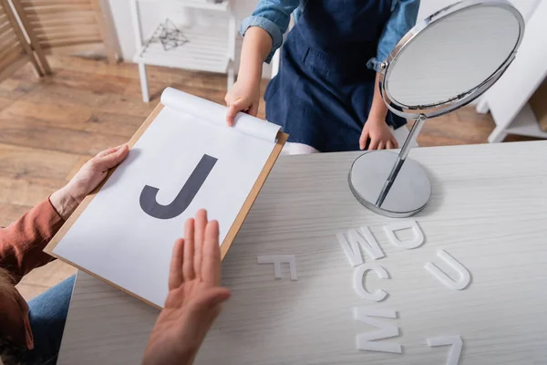 Vista ritagliata del logopedista e del bambino che indica una lettera negli appunti vicino allo specchio in classe — Foto stock