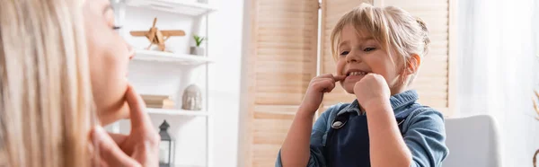 Girl pointing at mouth near speech therapist during lesson in classroom, banner — стоковое фото