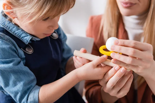 Terapeuta del habla borrosa sosteniendo entrenador de músculos respiratorios cerca de un niño en el aula - foto de stock