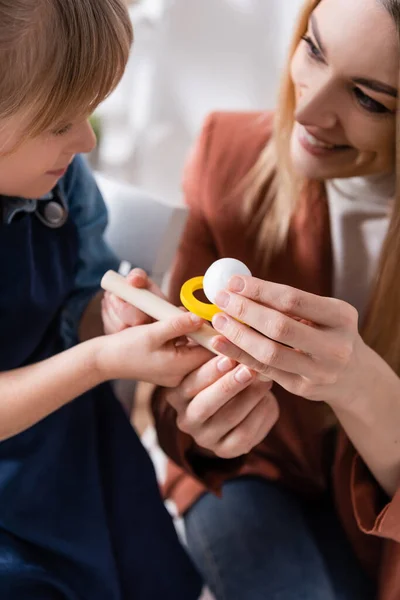 Smiling speech therapist holding respiratory muscle trainer near pupil in classroom — Stock Photo