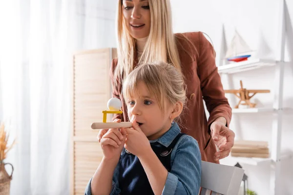 Child using respiratory muscle trainer near blurred speech therapist in classroom — стоковое фото