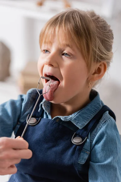 Speech therapist holding logopedic probe near kid sticking out tongue in classroom — Stock Photo