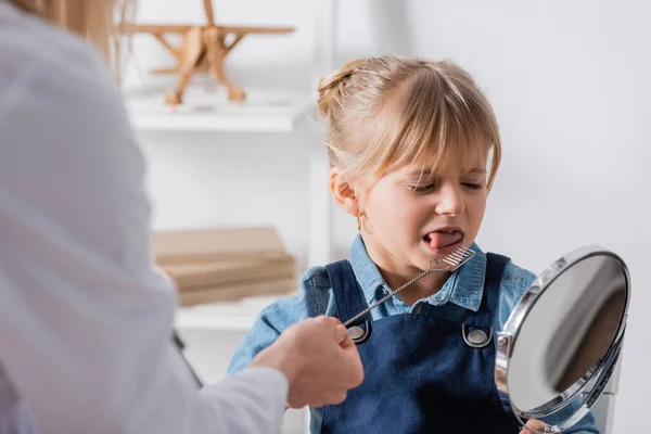 Niño sobresaliendo de la lengua cerca del terapeuta del habla con sonda logopédica y espejo en el aula — Stock Photo