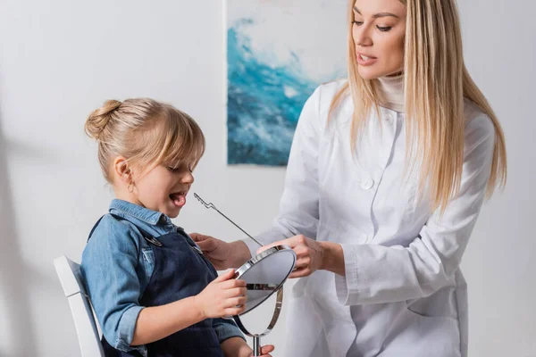 Terapeuta del habla en bata blanca sosteniendo sonda logopédica cerca de un niño con espejo en el aula - foto de stock