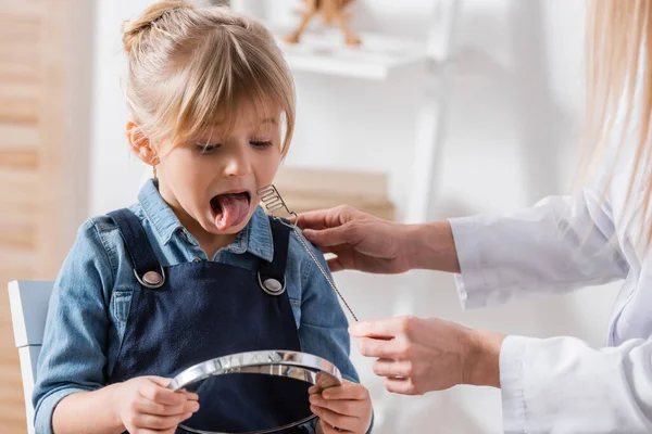 Chica sosteniendo espejo cerca de logopeda con sonda logopédica en el aula — Stock Photo