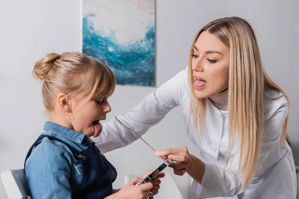 Speech therapist sticking out tongue and holding tool near girl with mirror in classroom — Stock Photo