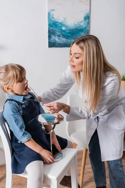 Terapeuta del habla en la capa blanca que sostiene la herramienta cerca de niña y espejo en el aula - foto de stock