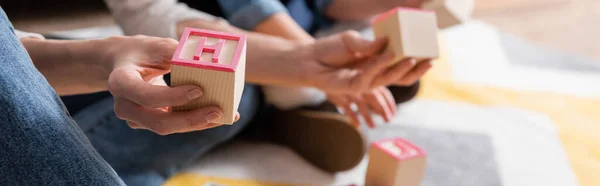 Vista recortada del logopedista sosteniendo un bloque de madera con carta cerca de un niño borroso en la sala de consulta, pancarta - foto de stock