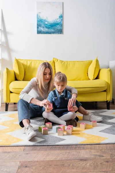 Kind hält Holzklötze mit Buchstaben neben Logopädin auf Teppich im Sprechzimmer — Stock Photo