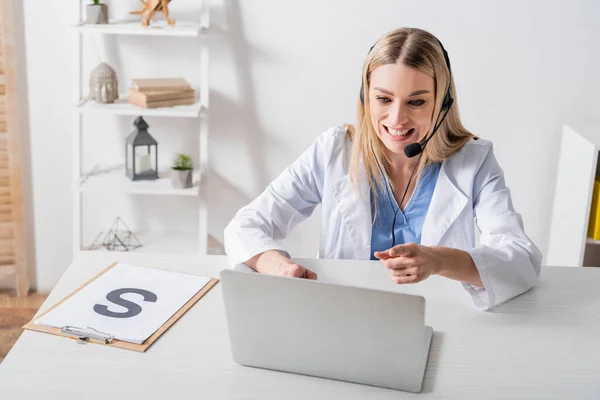 Smiling speech therapist in headset pointing at laptop during video call near clipboard in consulting room — Stock Photo