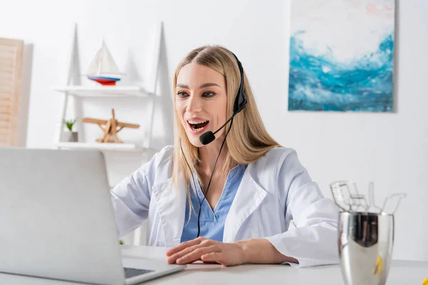 Cheerful speech therapist in headset having video call on blurred laptop near tools in consulting room — стоковое фото