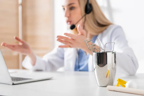 Tools and respiratory muscle trainer near blurred logopedist having video call on laptop in consulting room — стоковое фото