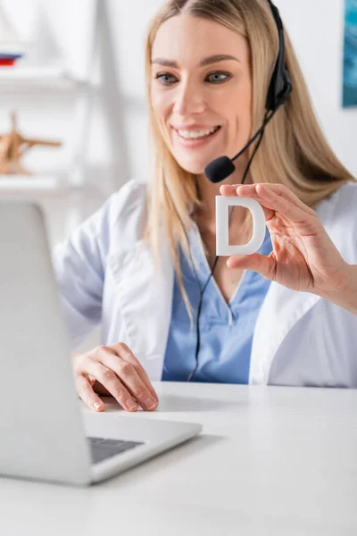 Smiling logopedist in headset holding letter near laptop during video call in consulting room — Stock Photo