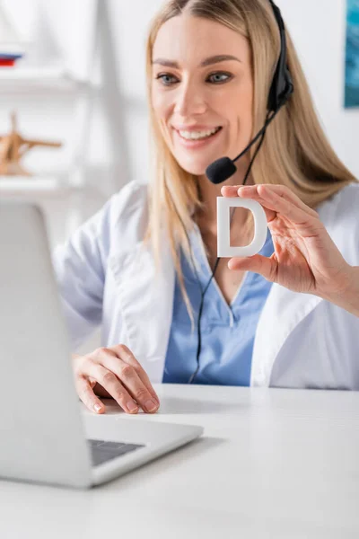 Logopedista sonriente en auriculares que sostiene la carta cerca de la computadora portátil durante la videollamada en la sala de consulta - foto de stock