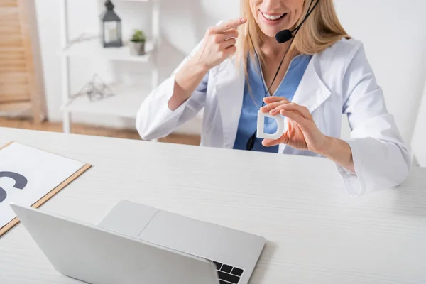 Vista ritagliata del logopedista sorridente in cuffia tenere lettera e puntare con il dito durante la videochiamata sul computer portatile in sala di consultazione — Foto stock