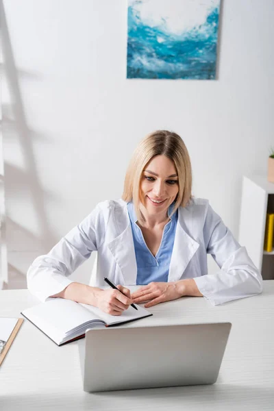 Positive speech therapist writing on notebook during video call on laptop in consulting room — Stock Photo
