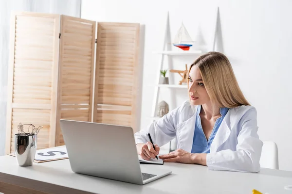 Speech therapist writing on notebook during video call on laptop in consulting room — стоковое фото