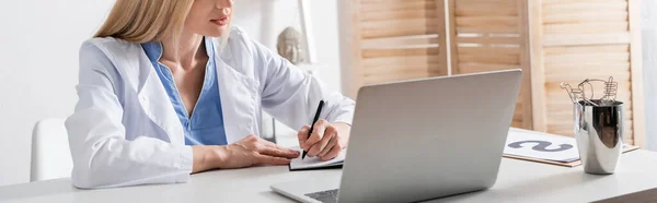 Vista recortada de la escritura del terapeuta del habla en el cuaderno cerca del ordenador portátil y portapapeles en la sala de consulta, pancarta - foto de stock